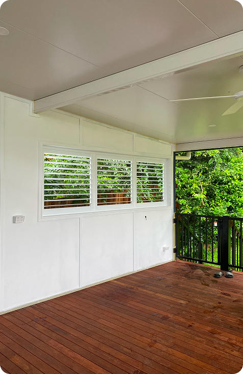 Covered patio with wooden floor, white walls, louvered window, and black railing. Green trees are visible outside through the window and the open space.