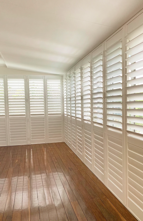 Bright room with wooden floor and large windows covered by white plantation shutters, allowing filtered sunlight to enter.