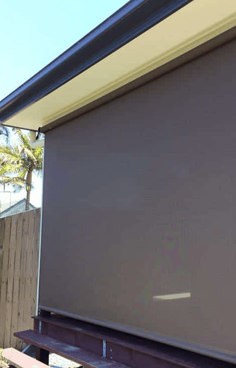 Outdoor roller blind fully extended, covering a patio area. Wooden fence, palm tree, and partial house roof visible in the background under clear sky.
