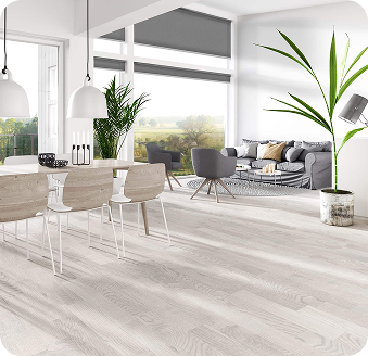 Modern open-plan living and dining area with light wood flooring, white furnishings, large windows, indoor plants, and a gray sofa set.