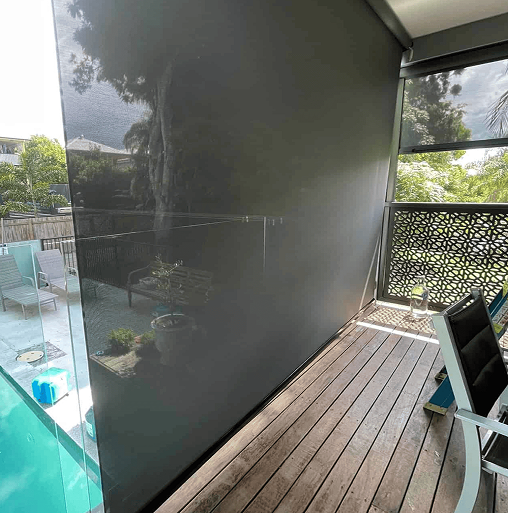 A semi-transparent outdoor roller blind covers one side of a balcony with wooden flooring, near a pool and outdoor furniture.
