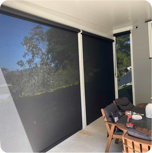 Outdoor covered patio with two large black roller blinds, a wooden table, four chairs, and some items on the table. Sunlight and trees are visible through the mesh blinds.