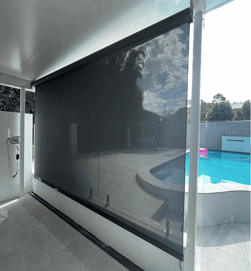 A black mesh outdoor blind covers a patio area next to a swimming pool, providing shade and partial privacy while allowing some light through.