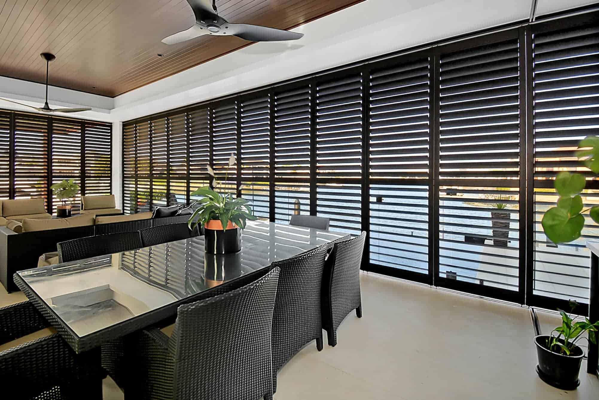 Modern enclosed patio with black louvered shutters, a glass dining table with wicker chairs, potted plants, ceiling fans, and a sitting area in the background.