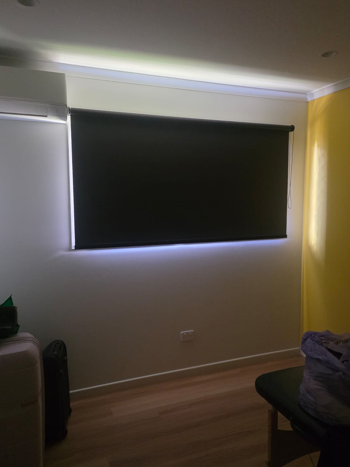 A dark roller blind covers a window in a small room with light yellow walls and wood flooring. Some light seeps from the edges of the blind.