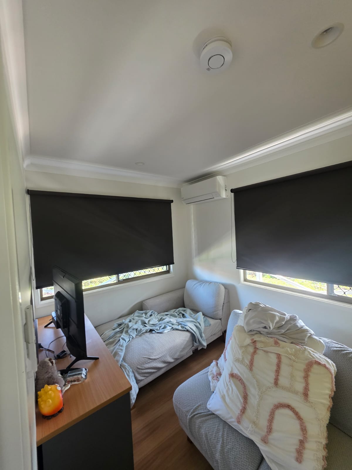 Small room with two couches, one with a blanket and pillow, a desk with a TV and plush toy, and windows covered by black blinds.