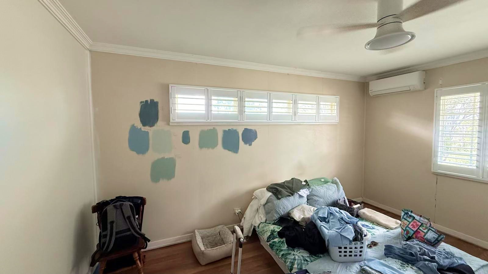 A bedroom with paint swatches on the wall, an unmade bed with clothes, a chair, a laundry basket, a pet bed, wood floor, and windows with white shutters.