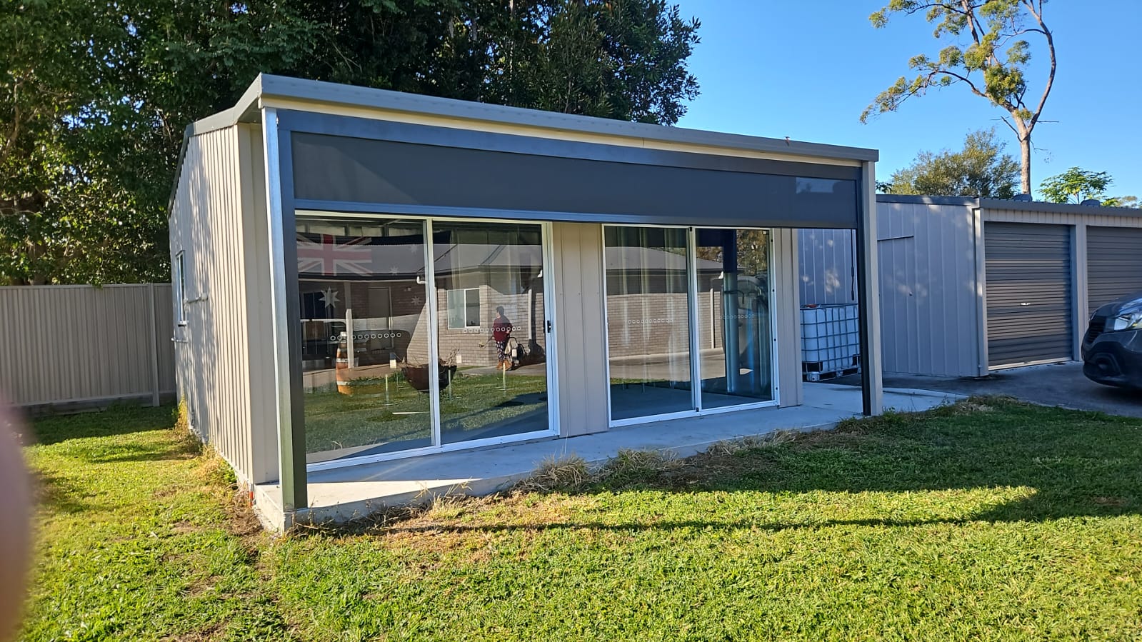 A small modern metal shed with large sliding glass doors sits on a concrete slab in a fenced backyard, next to a larger metal garage.