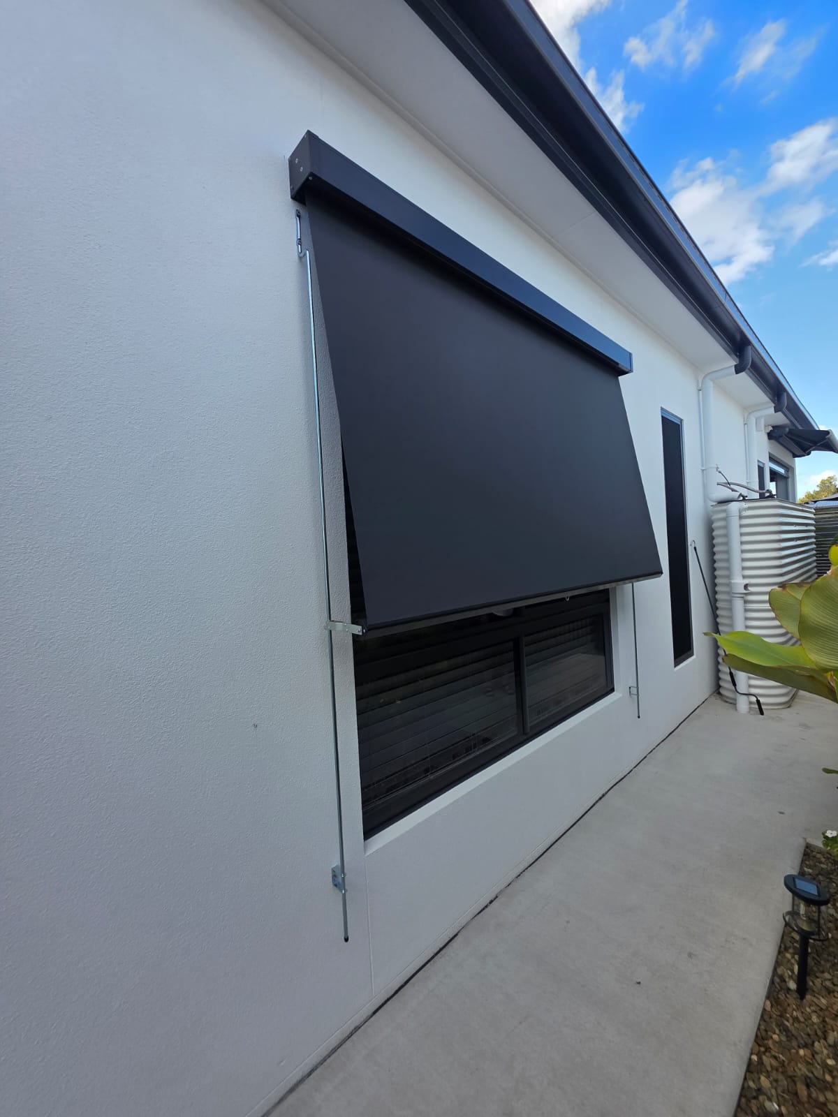Black outdoor awning partially extended over a window on a white building with a paved walkway beside it. Blue sky and clouds are visible in the background.