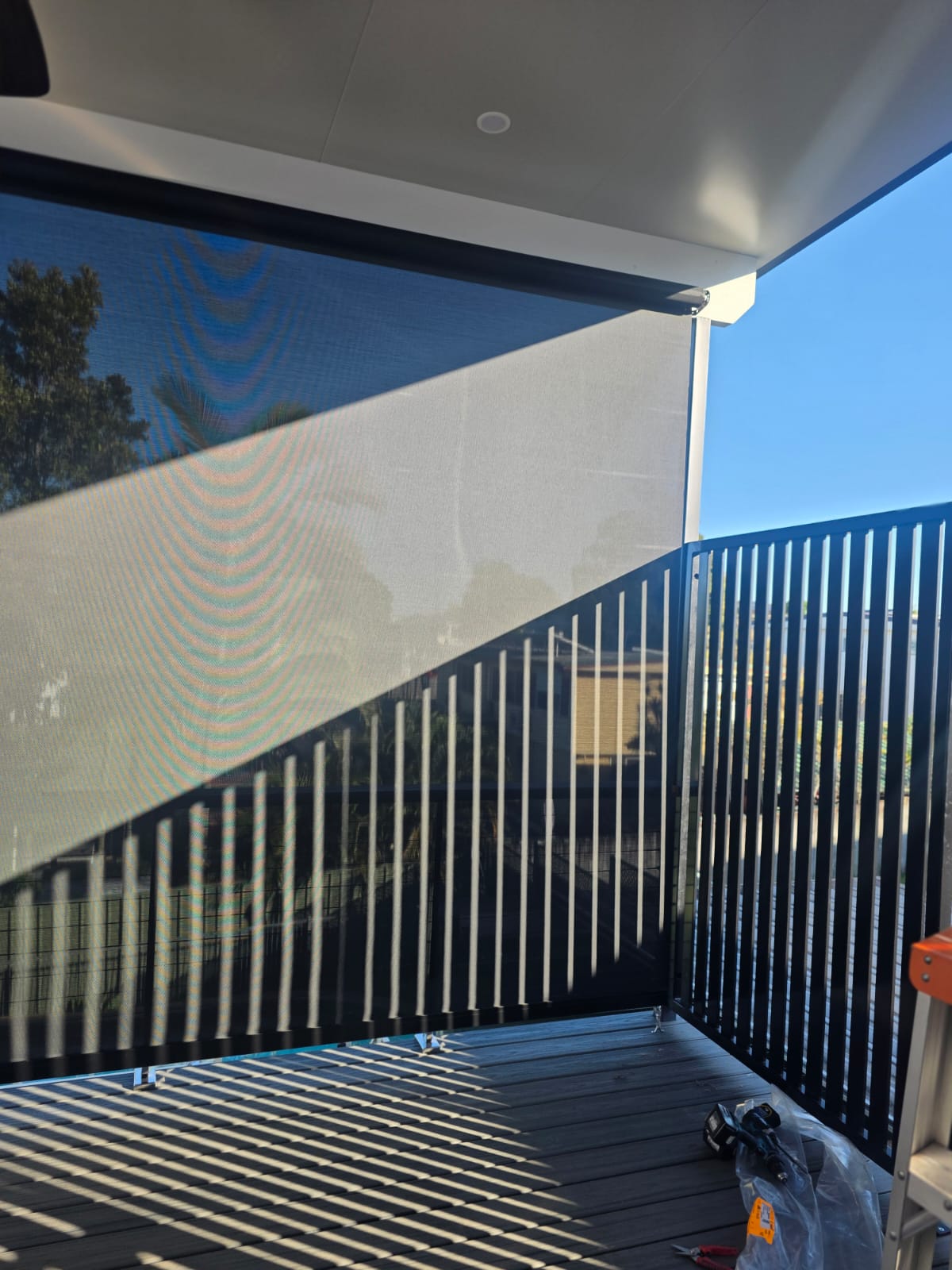 A balcony with a black mesh privacy screen and vertical metal railing casting striped shadows, with clear blue sky in the background.