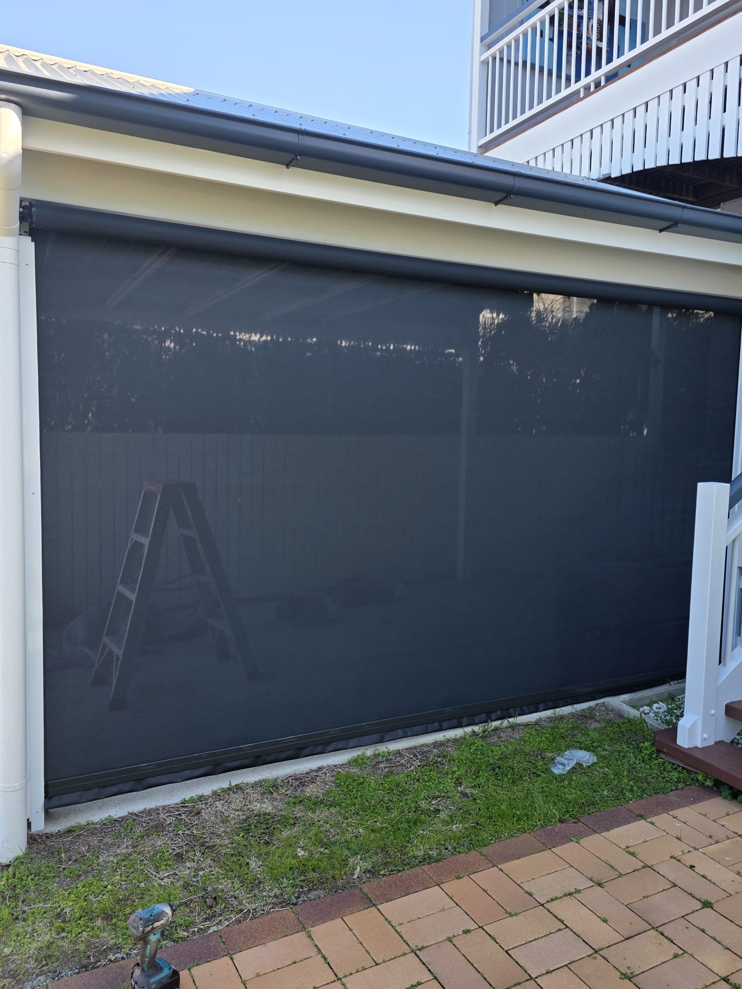 A black outdoor roller screen covers a patio area; a folded ladder is visible behind the screen, and a brick path runs alongside the grass in front.