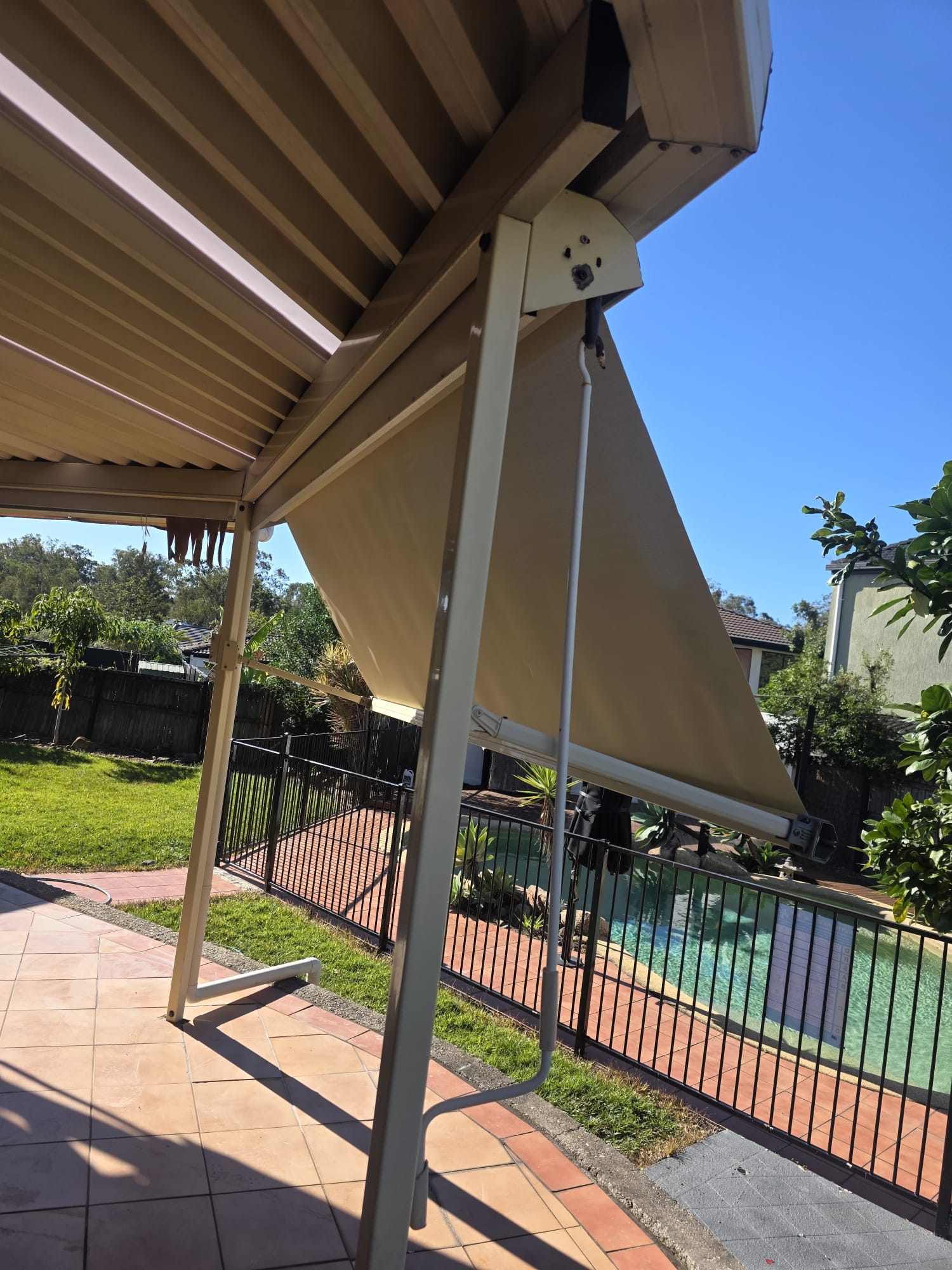 A retractable outdoor awning extends over a tiled patio next to a fenced backyard pool on a sunny day.
