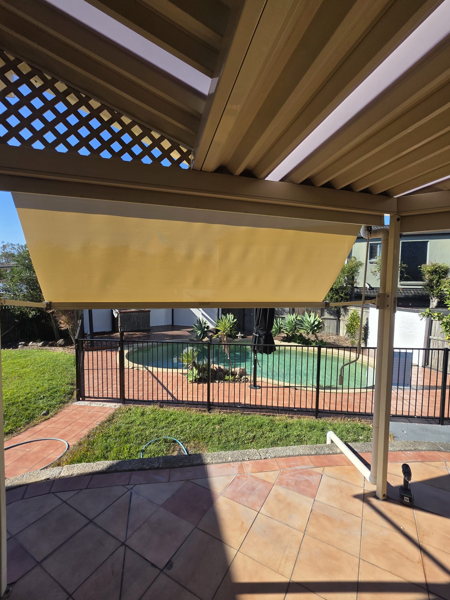 A yellow retractable shade is extended over a tiled patio, facing a fenced backyard with a swimming pool and plants, under a clear sky.