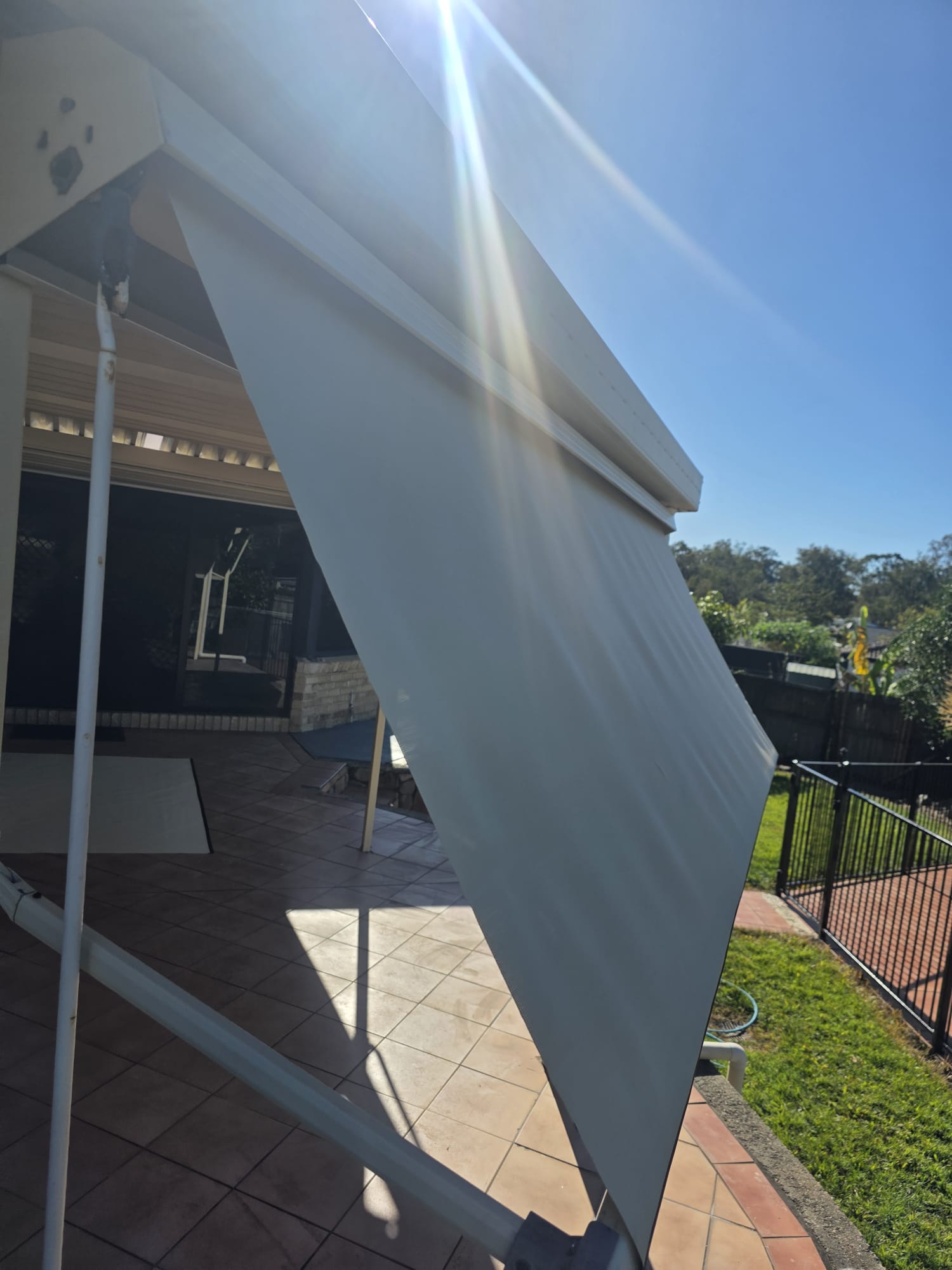 A white outdoor roller blind is partially extended over a tiled patio on a sunny day, with a garden and black fence in the background.