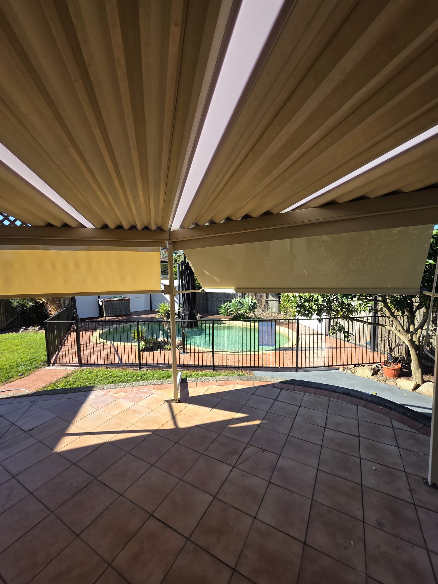 Covered patio with tiled floor, partially lowered outdoor blinds, and view of a fenced backyard with a circular pool and surrounding greenery.