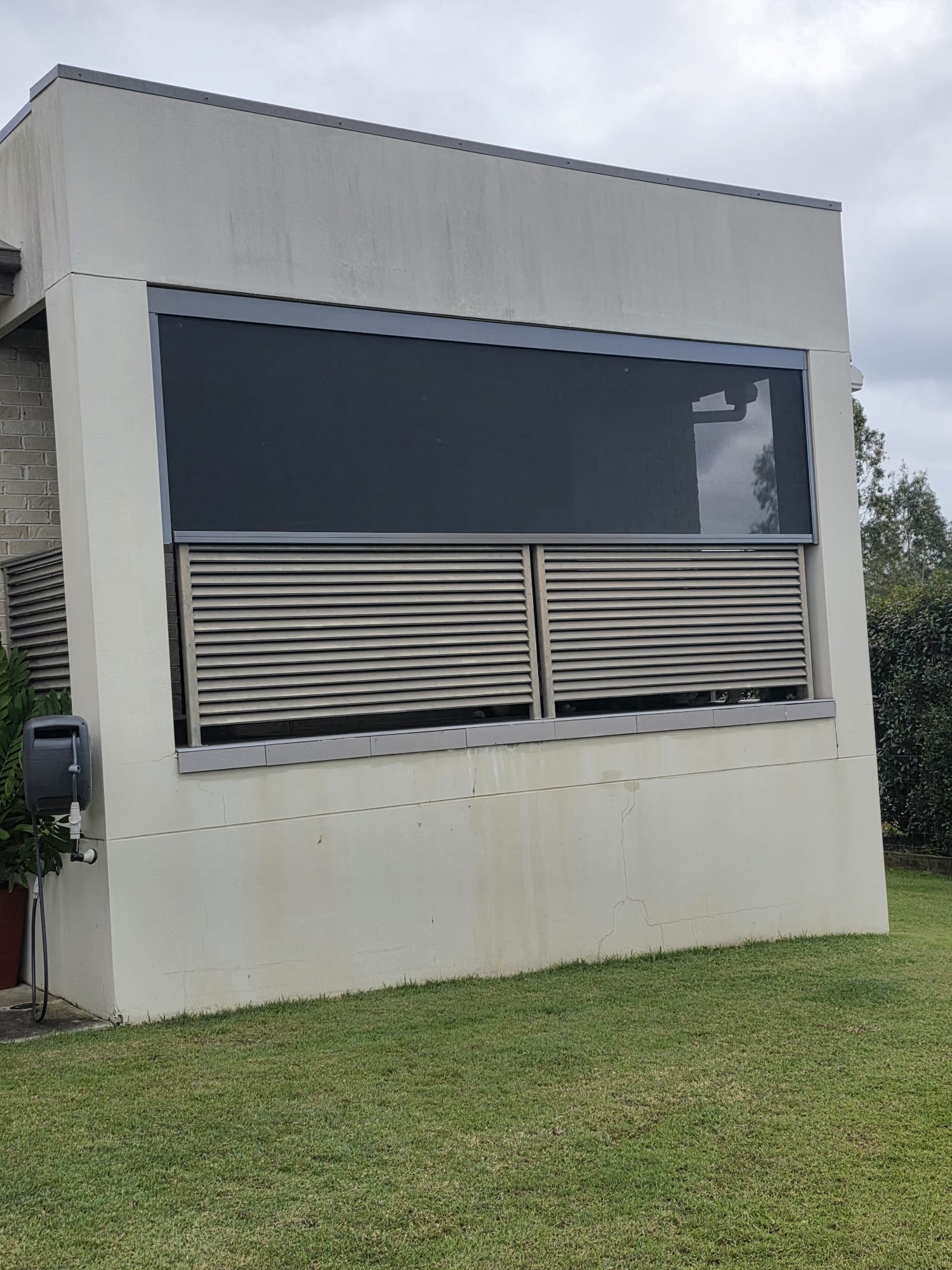 A modern building with a large tinted window and horizontal metal slats, white exterior walls, and a grassy lawn in the foreground under an overcast sky.