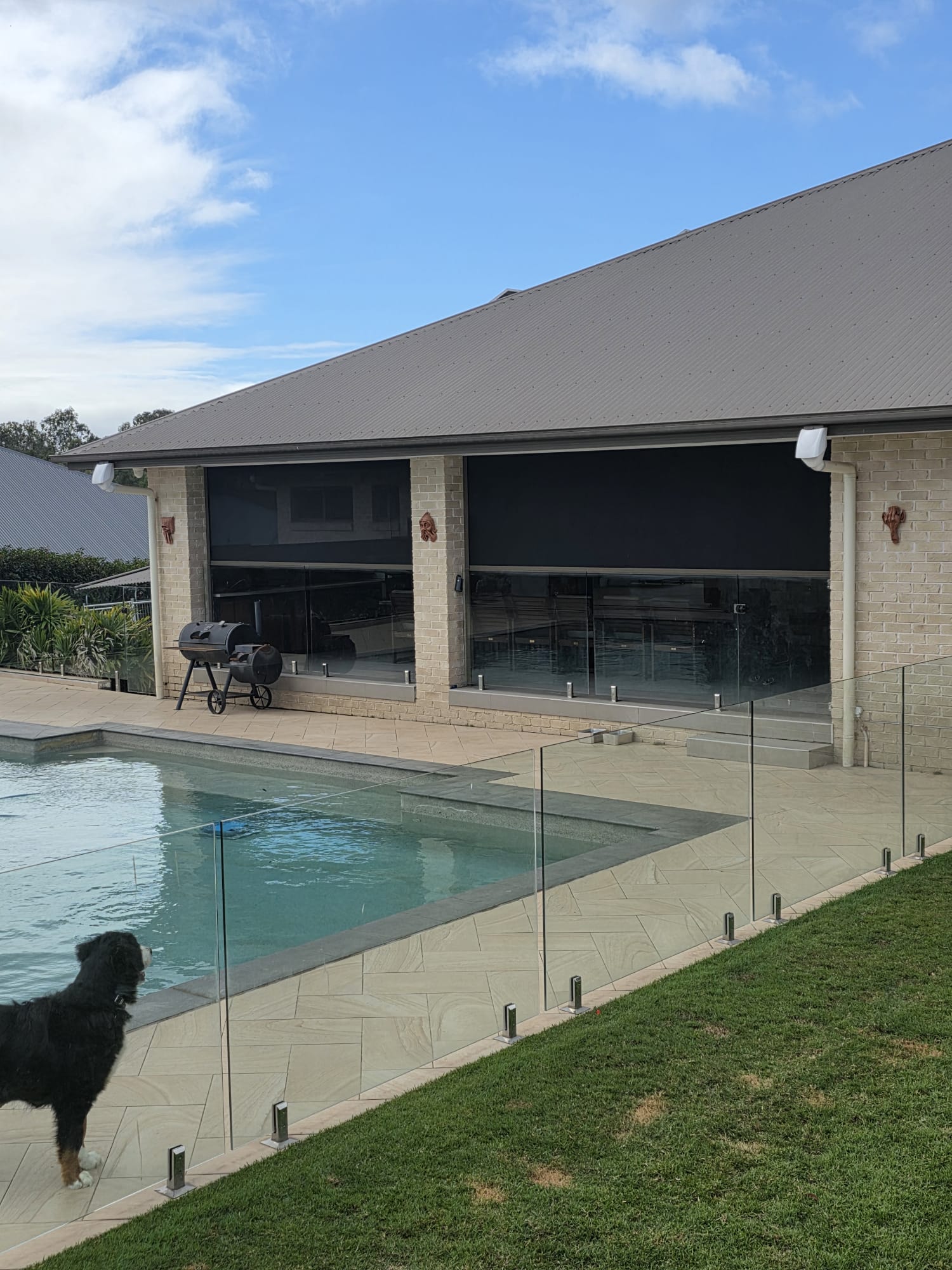 A covered patio with a grill and table overlooks a swimming pool, separated by a glass fence. A dog stands by the pool on the left.