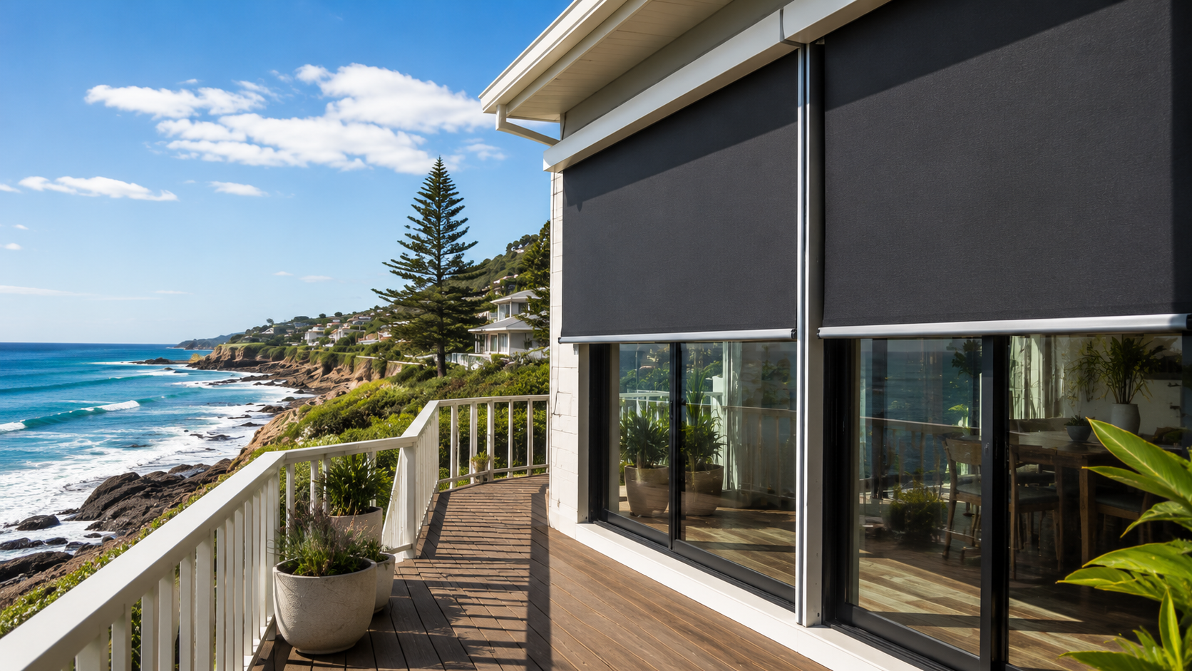 Modern house with large windows and black outdoor blinds on a wooden deck overlooking a coastal landscape with ocean and cliffs.