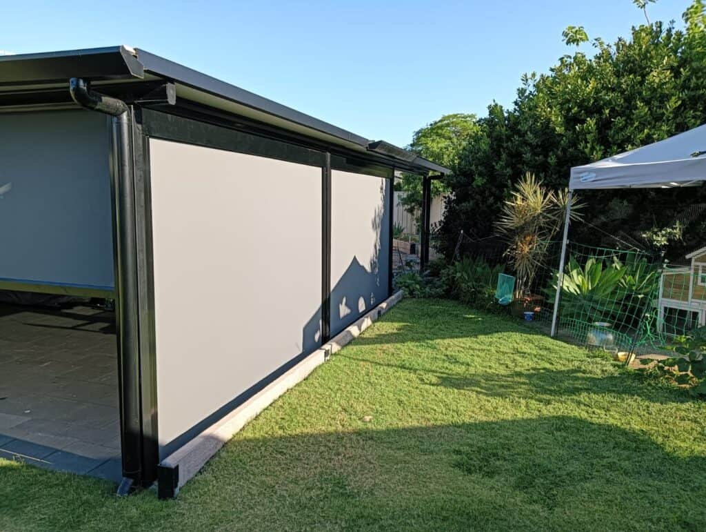 A modern outdoor patio with closed grey channel blinds beside a grassy yard with garden plants.