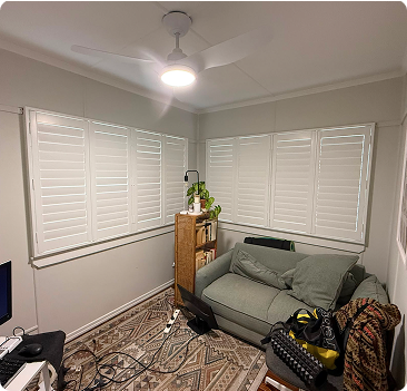 Small room with shuttered windows, a green couch, a shelf with a plant, a ceiling fan with light, a patterned rug, and various electronic cables on the floor.