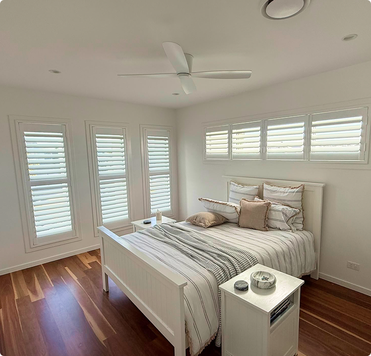 Bright bedroom with wooden floors, white walls, shuttered windows, a white bed with neutral bedding, some pillows, and two bedside tables with decor and a small candle.
