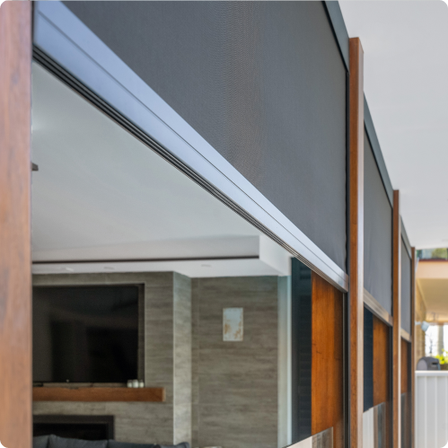 Retractable outdoor blinds partially lowered on a patio with wooden framing, overlooking a modern interior living area with a TV and neutral decor.