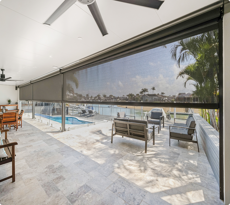 Covered patio with ceiling fans, lounge seating, and a dining table, overlooking a pool and waterfront, seen through partially lowered mesh sun shades.