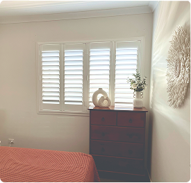 A bedroom with a red bedspread, a wooden dresser topped with decor, a plant, and a window with white shutters letting in daylight.