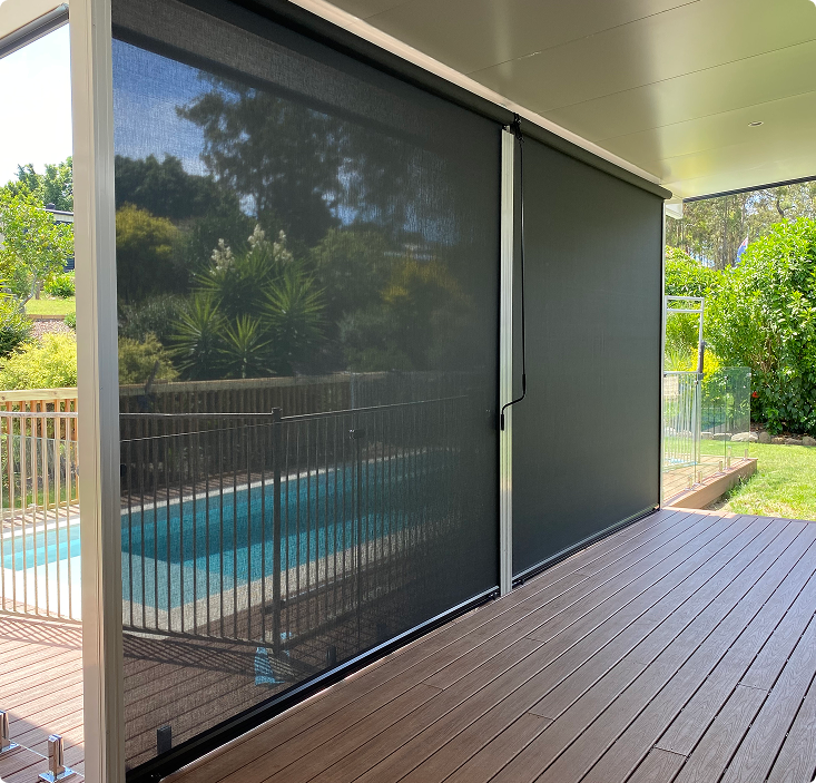 Black outdoor roller blinds cover a patio area with wooden flooring, partially shielding a view of a fenced-in backyard pool and green garden.
