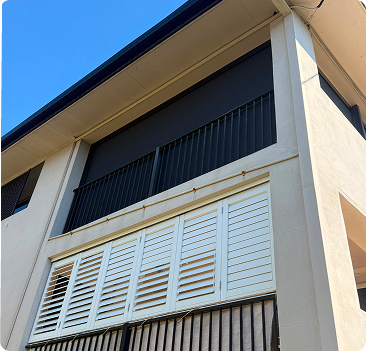 Two-story building with white louvered shutters on the lower balcony and black vertical bars on the upper level, viewed from below against a clear blue sky.
