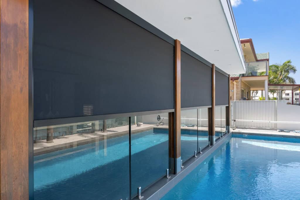Outdoor roller blinds partially cover glass panels next to a modern swimming pool under a covered patio, creating a stylish and comfortable retreat in Newport on a sunny day.