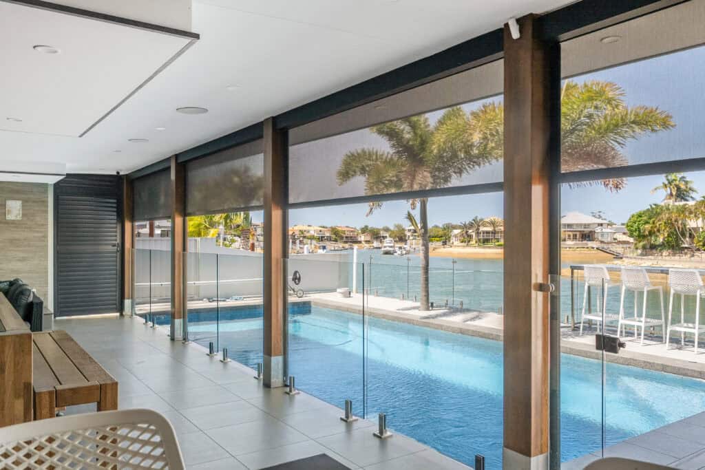 A covered patio with glass fencing overlooking a swimming pool and waterway in Cleveland, with palm trees and houses in the background.