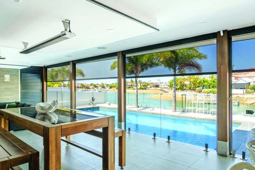 Modern indoor-outdoor space in Redcliffe with glass doors, wooden dining table, pool, palm trees, and a waterfront view under a clear sky.
