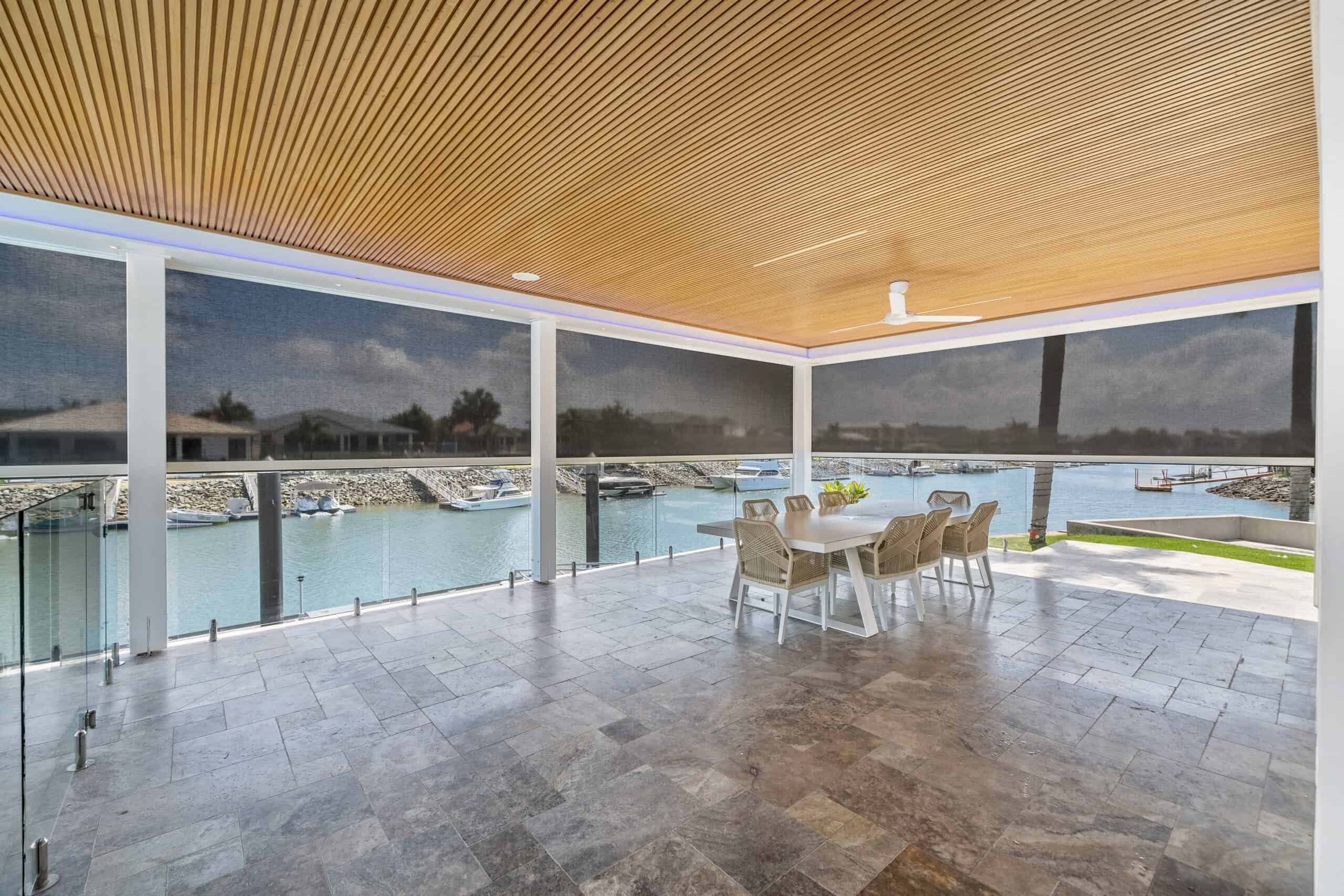 Covered patio with tiled floor, wooden ceiling, ceiling fan, dining table for six, glass railing, and a scenic waterfront view.