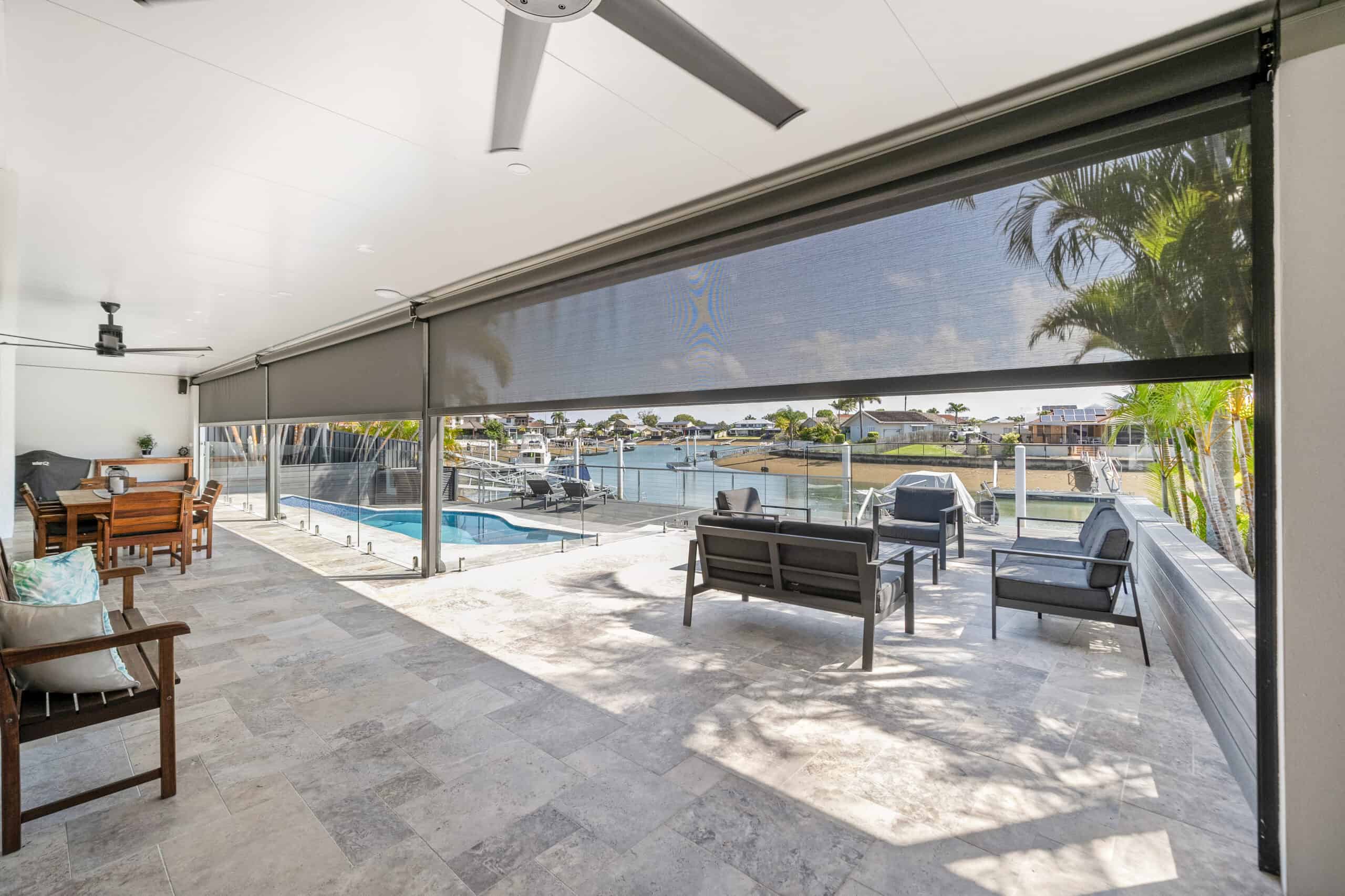 Covered patio with outdoor seating and ceiling fans, offering views of the pool and waterfront Redcliffe homes through glass fencing and roll-down shades.