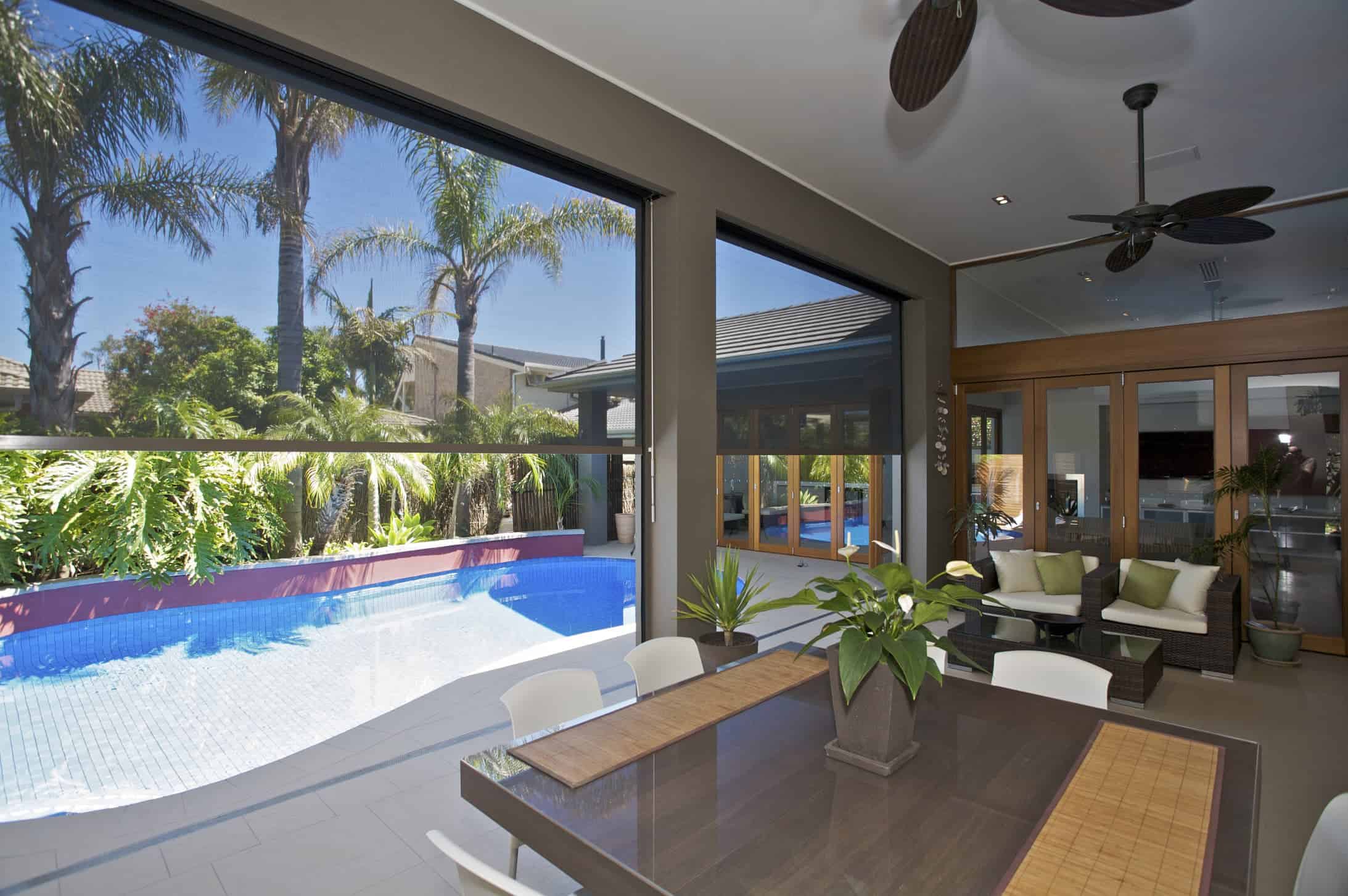 Modern covered patio with dining table and potted plants overlooks a backyard swimming pool surrounded by palm trees and outdoor seating.