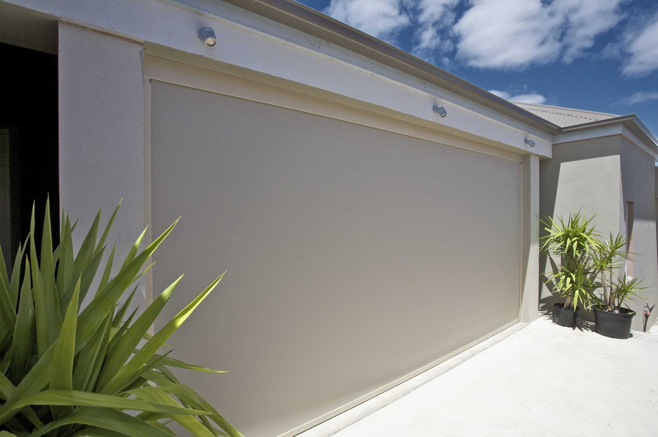 A modern house exterior with a large beige roller shutter door, potted plants, and a partly cloudy blue sky above.