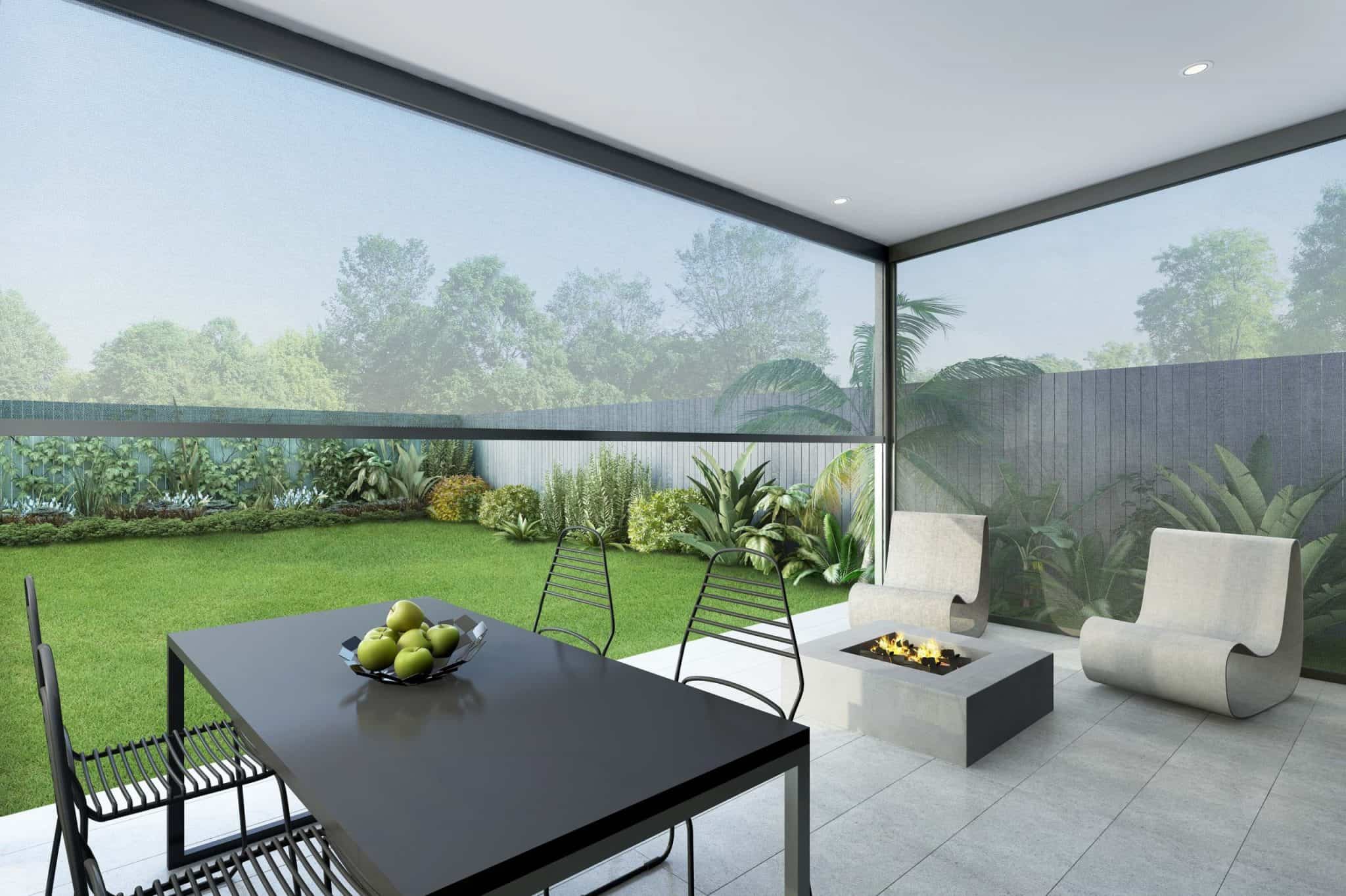 Modern outdoor patio with two white chairs, a black dining table with four chairs, a bowl of green apples, and a fire pit, overlooking a green garden with trees and plants.