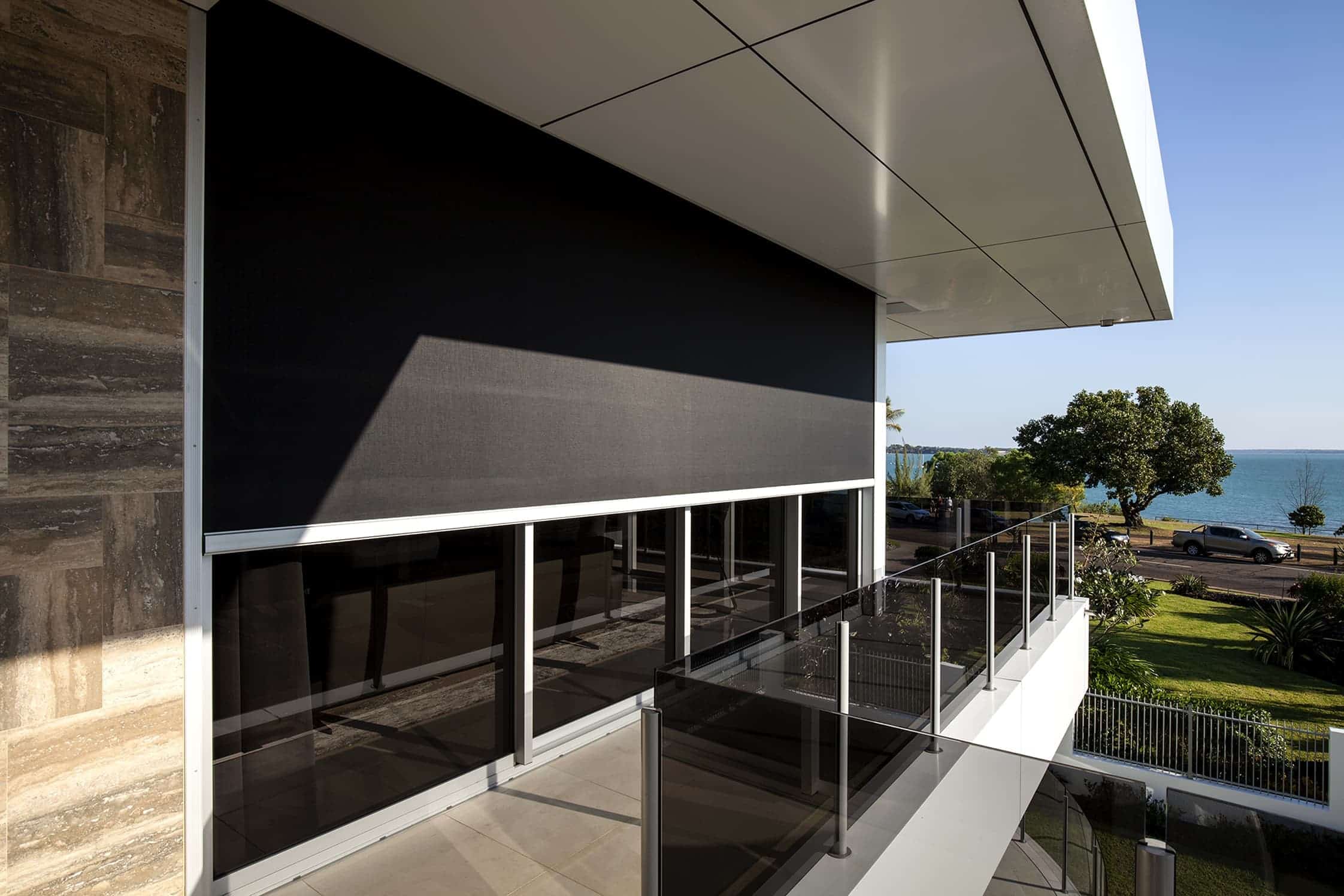 Modern building balcony with glass railing and a large black retractable screen overlooking a green lawn and trees, with a view of water and sky in the background.