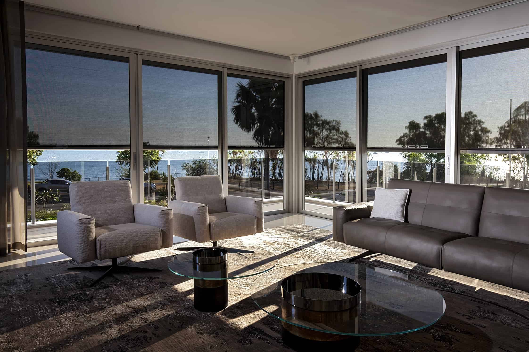 Modern living room with gray furniture, glass coffee tables, and large windows showing a view of trees, road, and ocean outside.