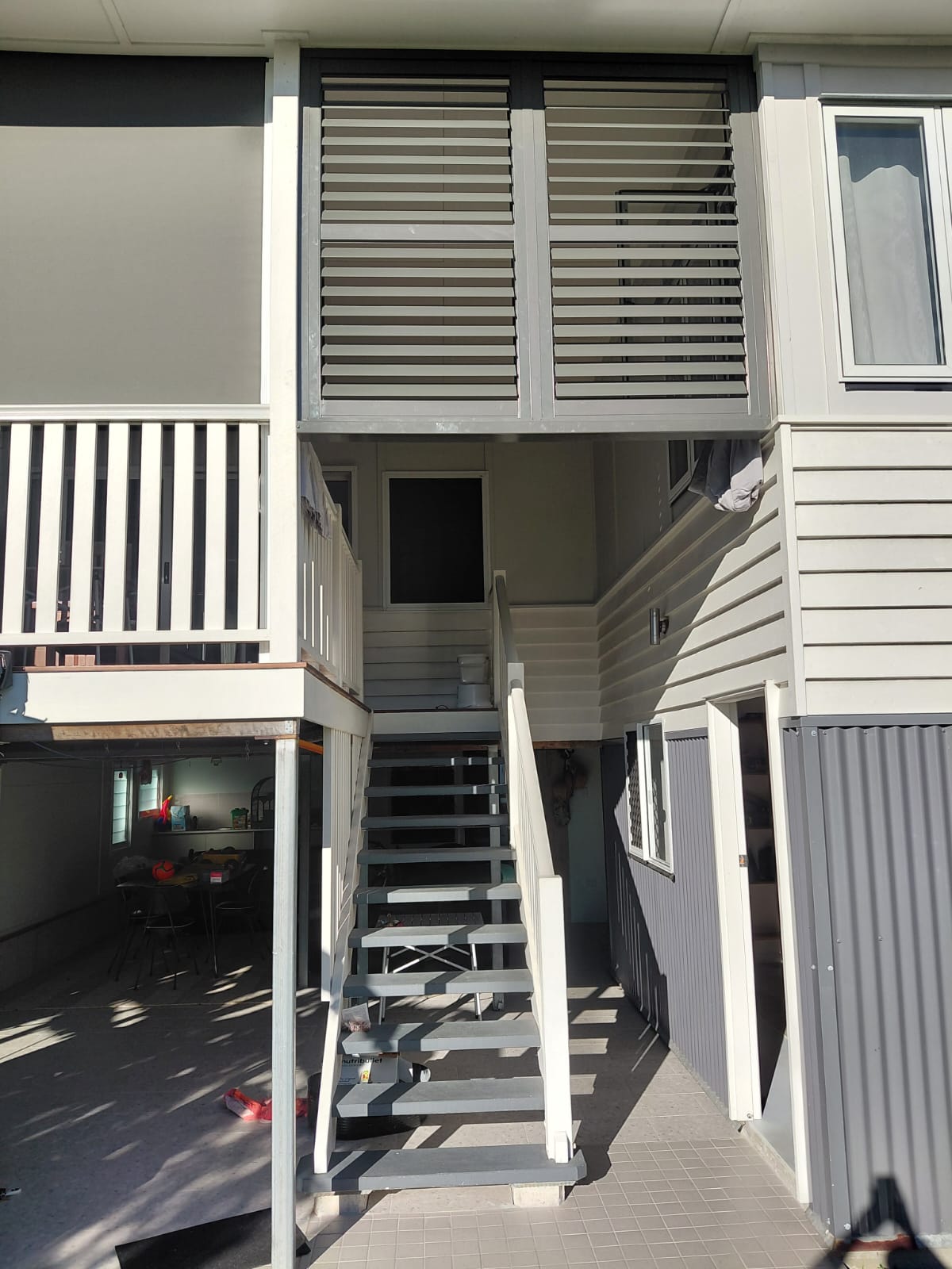 Exterior view of a house with an open staircase leading to an upper level, louvered windows, and a shaded ground floor area with a table and chairs visible.