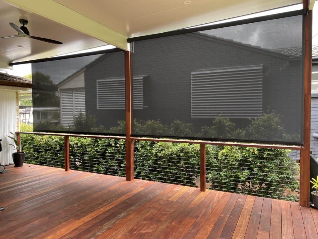 Outdoor patio with a timber deck, ceiling fan and two roll-down mesh blinds partially lowered over the view of the garden.