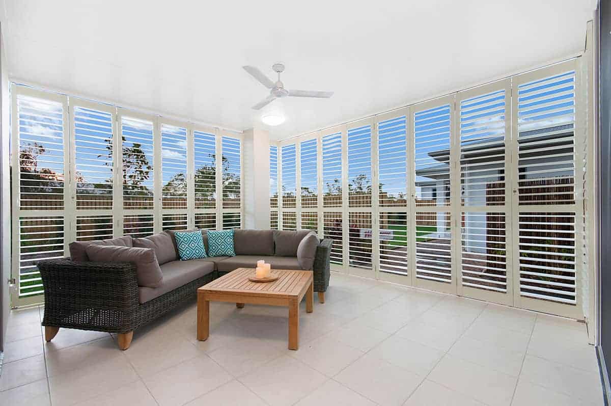 Enclosed patio with white tile floor, wicker sofa with blue and white cushions, wooden coffee table, ceiling fan, and floor-to-ceiling windows with white shutters.