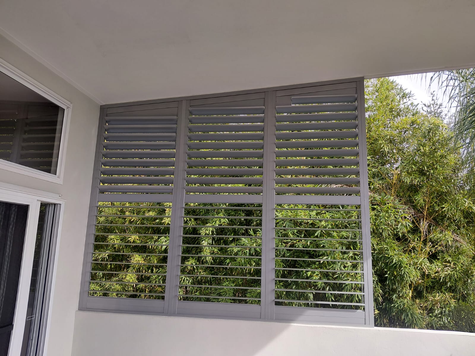 Gray metal louvered panels are installed on a balcony, partially shading the area while green bamboo plants are visible outside.