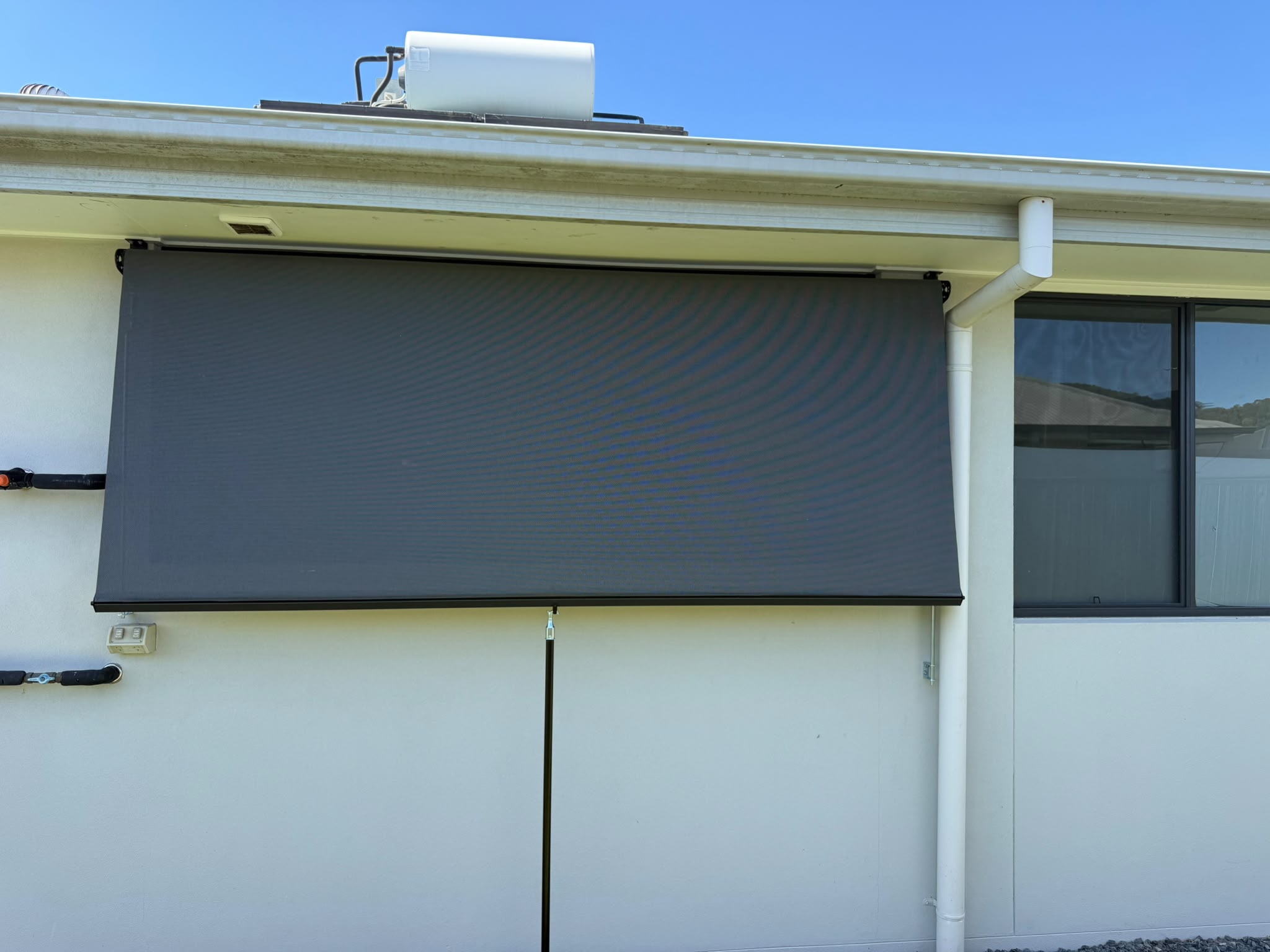 A black outdoor roller shade is mounted on the exterior wall of a building below the roof, next to a window, with a clear blue sky above.