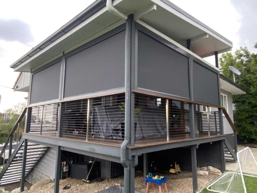 A modern elevated Morayfield house with a covered balcony enclosed by dark privacy screens and metal railings, featuring gravel and storage space underneath.