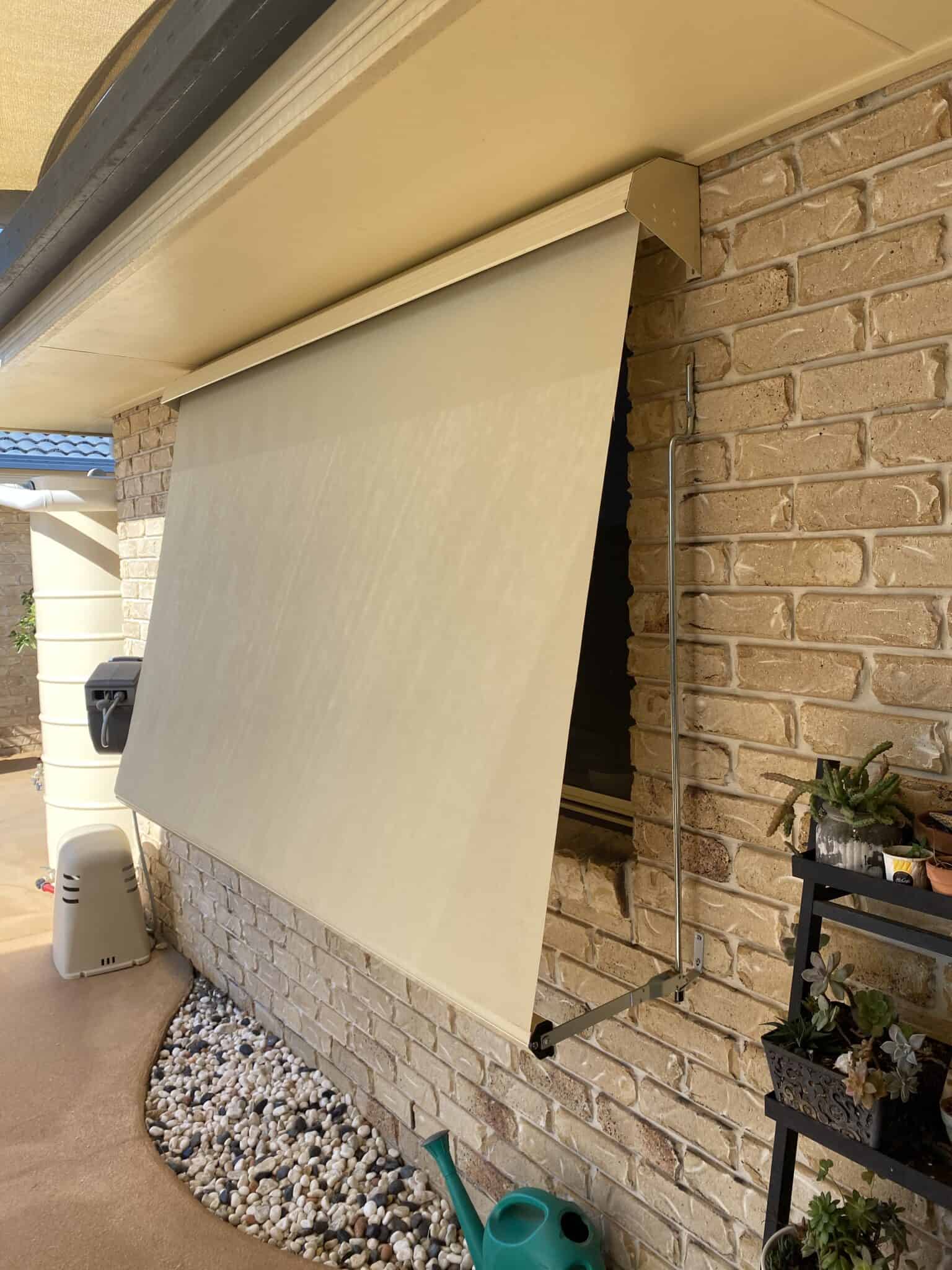 A beige outdoor roller shade partially covers a window on a brick wall, with a rock garden, watering can, and potted plants nearby.