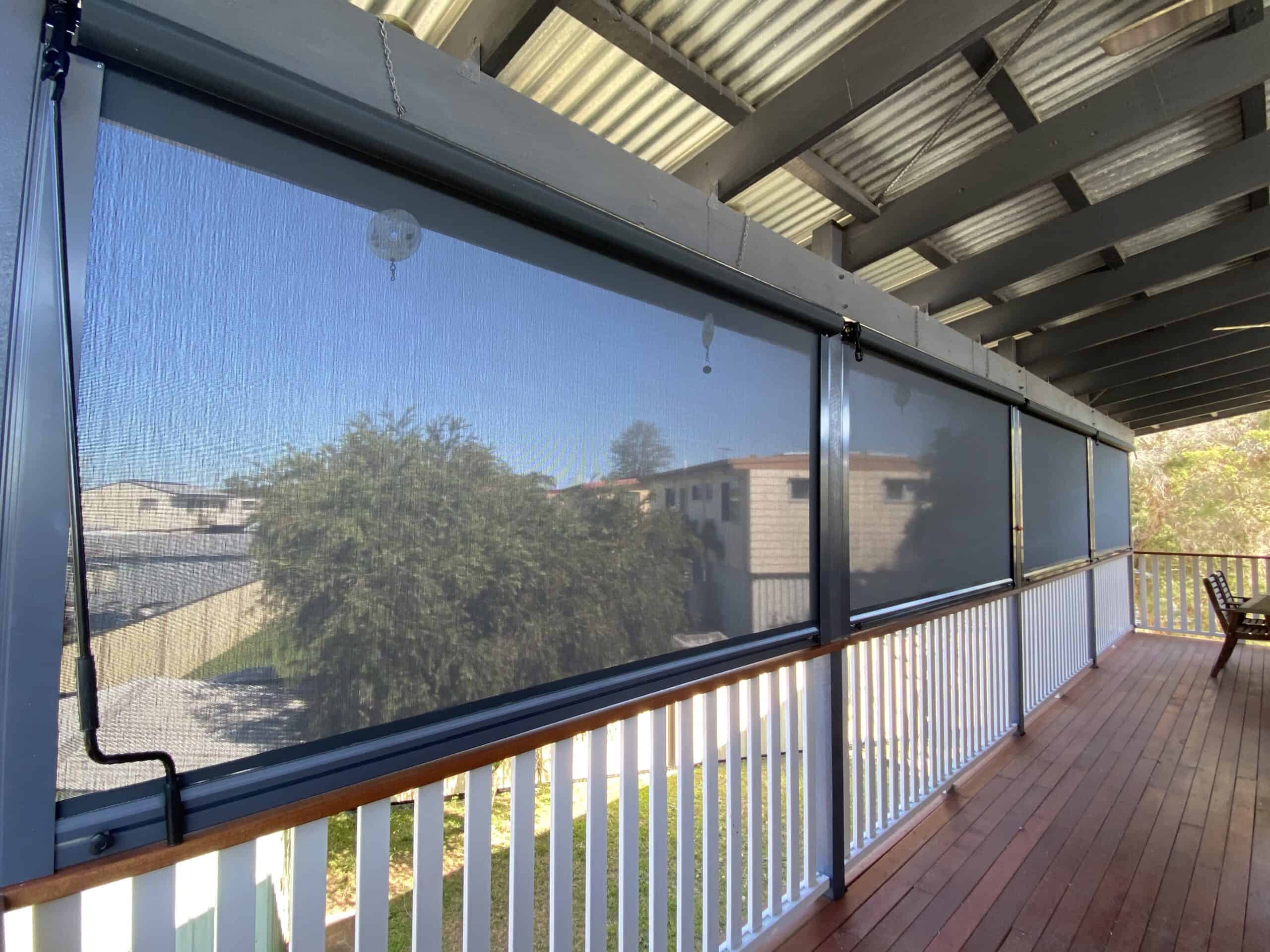 Outdoor covered deck with wooden floor and white railing, fitted with roll-down mesh screens along the side, partially revealing houses and trees in Morayfield.