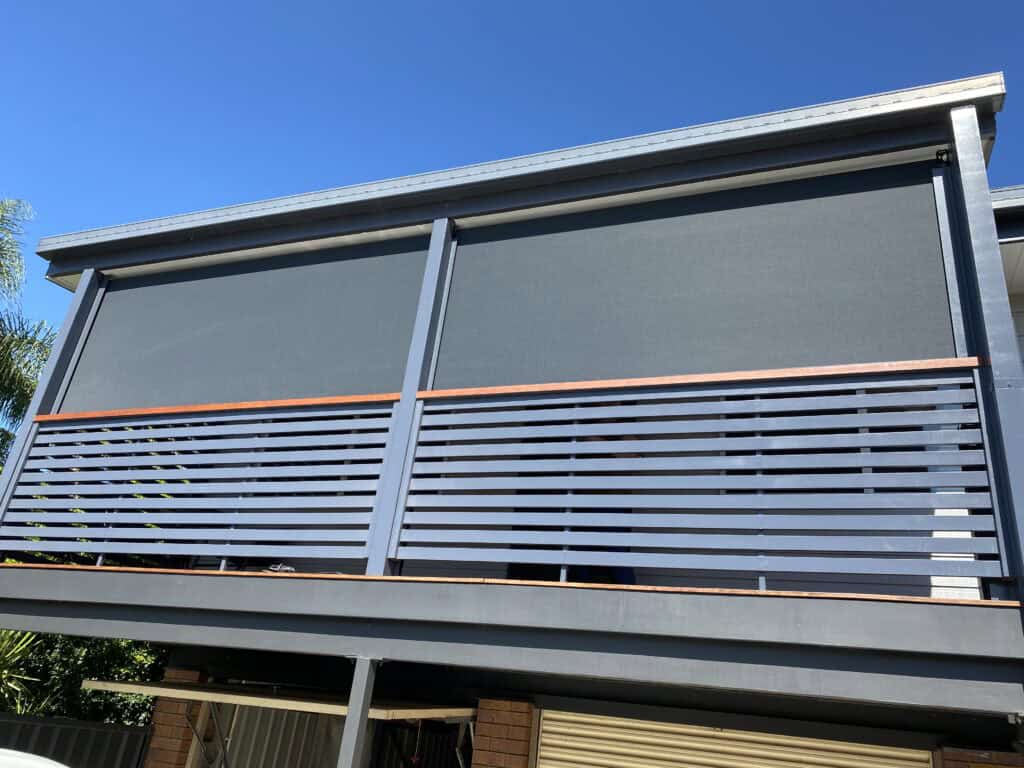 A modern balcony with two large outdoor roller blinds fully lowered, horizontal metal railing, and a clear blue sky in the background.