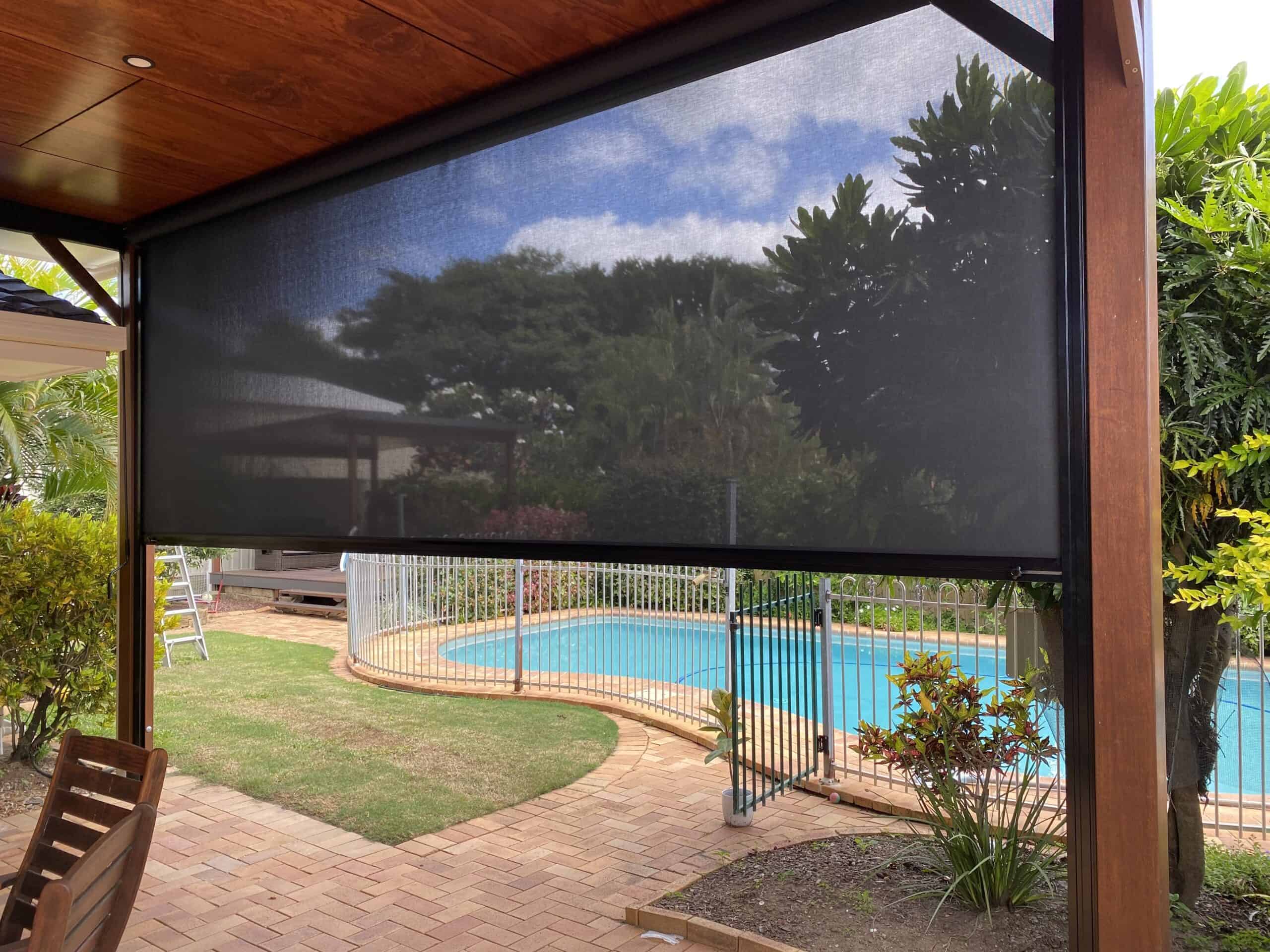 A backyard patio in Burpengary with a black mesh shade, overlooking a fenced swimming pool, brick paving, and lush green plants under a partly cloudy sky.
